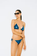 Woman modeling metallic blue bikini with gold chain straps and black sunglasses against white background